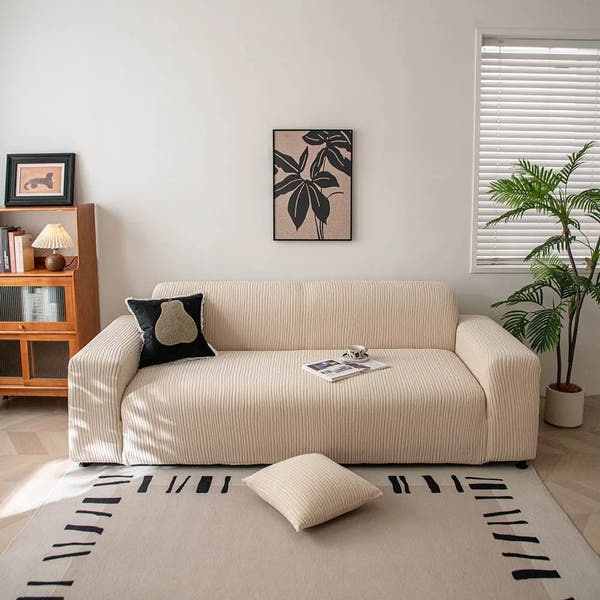 Linen beige corduroy stretch sofa cover in a bright living room with framed art and plant
