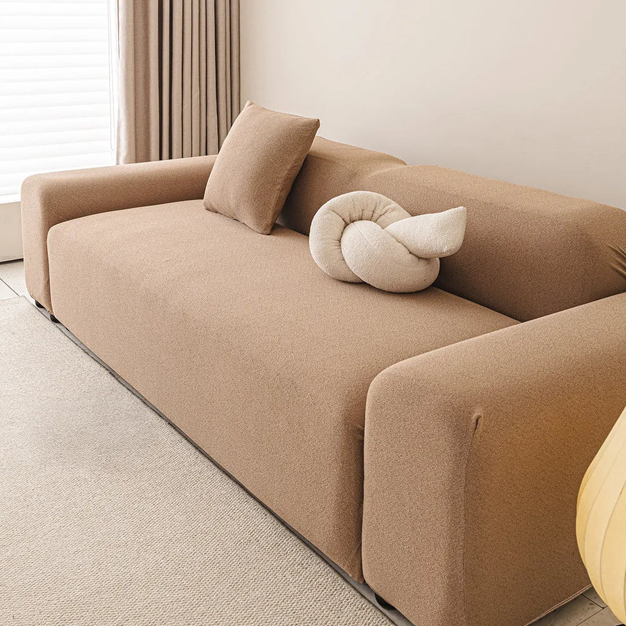 Sunlit living room sofa in caramel sand smooth cover, viewed from the right with a knot cushion.