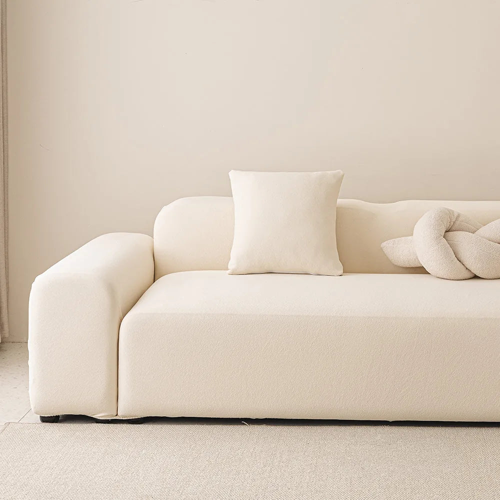 Cream smooth stretch sofa cover styled on a corner sofa with a cushion and knot pillow