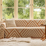Geometric rust chevron throw covering a sofa by a sunlit window with ivory cushion and side table