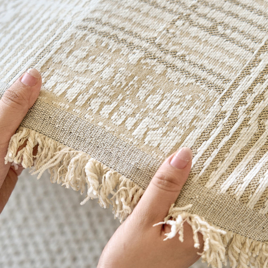 Hands holding the sand beige mosaic fabric showing woven texture and fringed edge